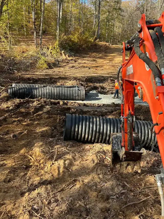 Orange Kubota excavator arm over black corrugated culvert pipe on a cleared woods road