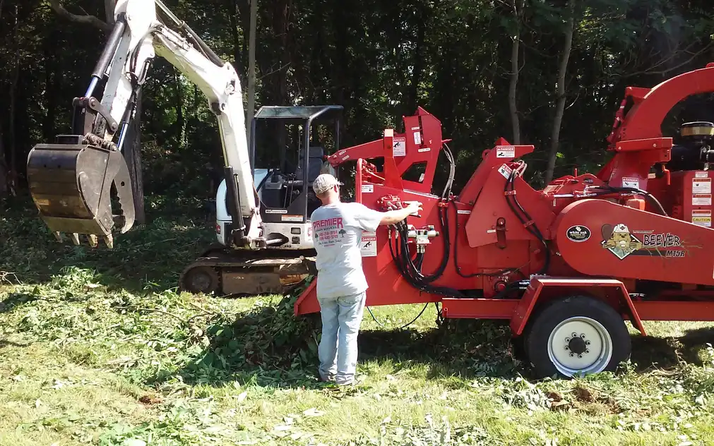 Premier crew member feeding limbs into a red Morbark Beever M15R chipper beside a grapple excavator